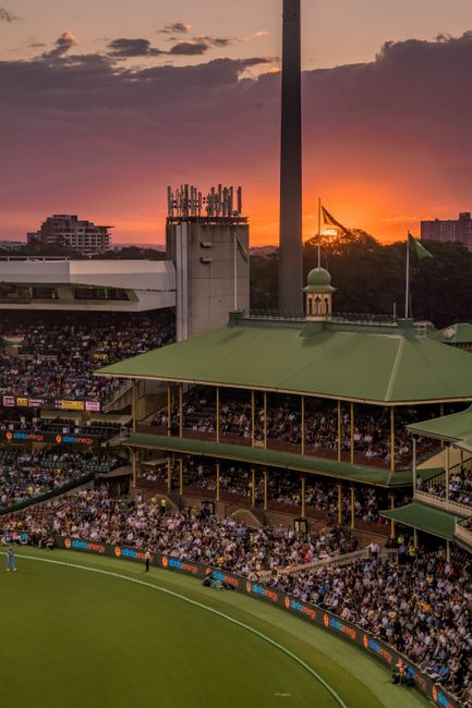 The image shows a view of a large cricket stadium during sunset, with a crowd of spectators seated under multiple green, tiered roofs supported by wooden and metal structures. Behind the stadium, a ci